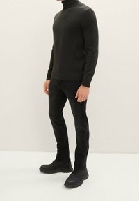 Mand iført en sort højhalset sweater, sorte jeans og sorte snørestøvler, står mod en ensfarvet lys baggrund.