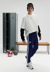 Balta pārizmēra T-krekls pār melnas garām piedurknēm kreklu, tumši zili sporta bikses ar svītrām un baltas sporta kurpes. Fonā redzama skapis un sporta bumbas.