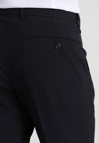 Dunkelblaue Hose mit glatter Textur, ausgestattet mit einer Gesäßtasche und einem schwarzen Knopfverschluss. Maßgeschneiderter Schnitt mit minimalistischen Akzenten.