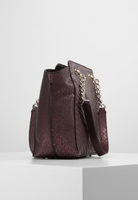 Bolso de mano burdeos con patrón en relieve y asas de cadena plateadas, exhibido sobre un pedestal blanco contra un fondo liso.