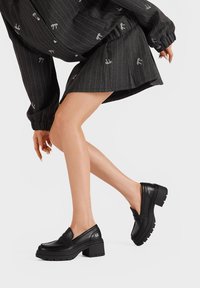 Scarpe in pelle nera con tacco grosso e design slip-on. Presentano una parte anteriore cucita e una suola ribattuta per aderenza e stabilità.