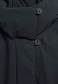 Zwarte trenchcoat met hoge kraag, diagonale geborduurde textuur en een ronde zwarte knoopsluiting. Glad materiaal met naden.