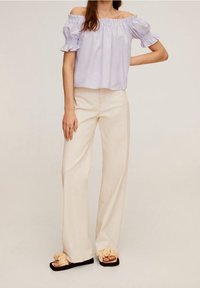 Top viola chiaro a spalla scoperta con maniche a volant, abbinato a pantaloni beige a gamba larga e sandali neri con dettagli a volant.