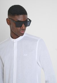 Camicia di lino bianca con colletto e chiusura frontale a bottoni, con un piccolo logo ricamato. Occhiali da sole neri oversize con montatura angolare.