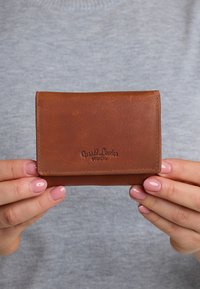 Rudas odinis piniginė su lygiu pavir�šiumi, sulankstomu dizainu ir iškilniu logotipu. Laikoma rankose prieš pilką foną.