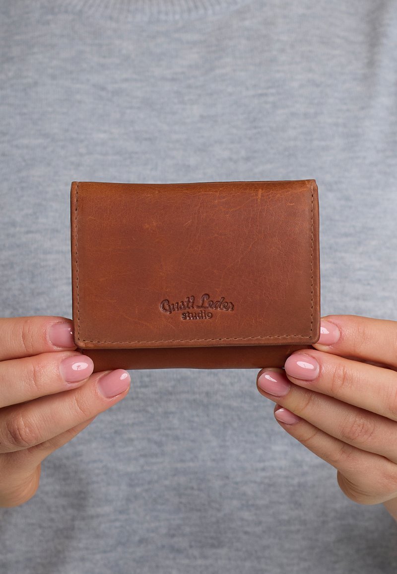 Rudas odinis piniginė su lygiu paviršiumi, sulankstomu dizainu ir iškilniu logotipu. Laikoma rankose prieš pilką foną.