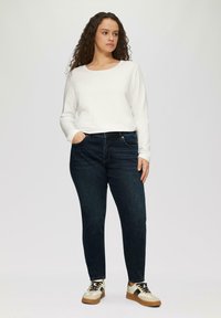 Biała koszula z długim rękawem w połączeniu z ciemnoniebieskimi dżinsami. Model nosi beżowe sneakersy z czarnymi akcentami i wzorzystą podeszwą. Prosty design.