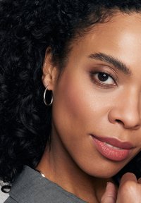 Srebrna kolczyki w kształcie kół, okrągłe, średniej wielkości. Kręcone czarne włosy, naturalny odcień skóry, neutralna szminka, subtelny makijaż oczu, szary T-shirt.