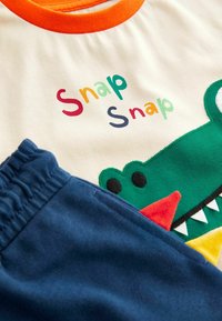 Short-sleeve cream t-shirt with orange collar, featuring colorful "Snap Snap" text and green crocodile design; paired with navy shorts.