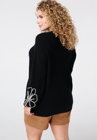 Zwarte gebreide trui met brede mouwen, voorzien van een witte geborduurde bloemdetails op de linkerarm, gecombineerd met tan shorts.