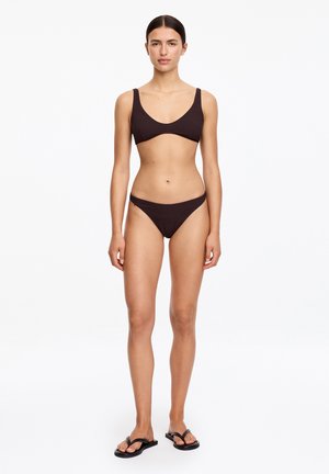 Femme debout, de face, portant un bikini texturé brun foncé et des tongs noires sur un fond blanc uni.