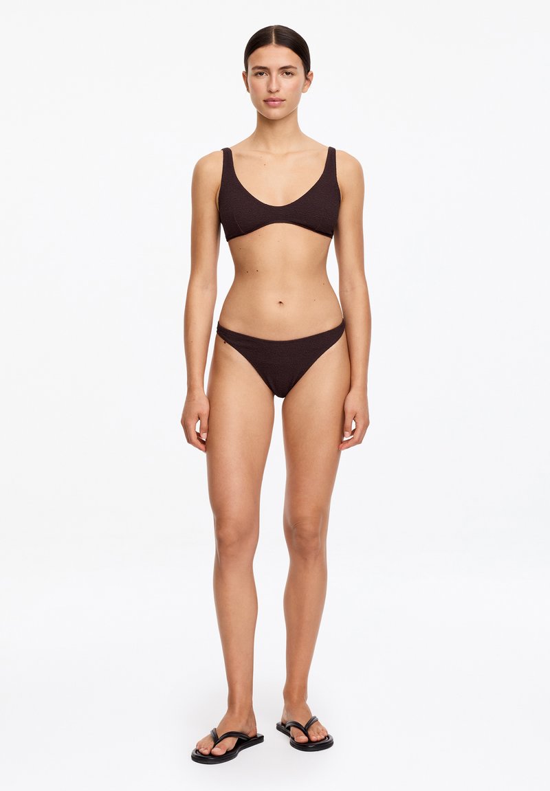 Femme debout, de face, portant un bikini texturé brun foncé et des tongs noires sur un fond blanc uni.