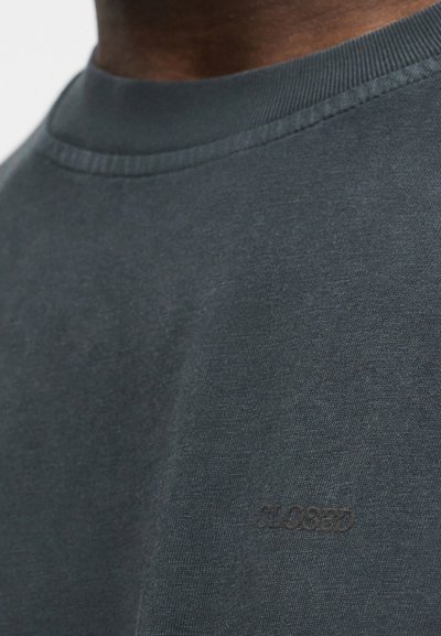 Κοντινό πλάνο σκούρου γκρι μπλουζιού με λαιμόκοψη τύπου crewneck, με διακριτικό ανάγλυφο λογότυπο «CLOSED» στο στήθος, φορεμένο από άτομο με σκουρόχρωμο δέρμα.