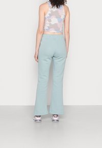 Žena oblečená v pastelovém abstraktním bezrukávovém topu, světle modrých rozšířených kalhotách a růžových a bílých teniskách, stojící čelem k bílé zdi.