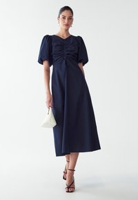 Mørkeblå midi-kjole med pufærmer og sammenkrøllede detaljer foran, parret med en hvid trekantet håndtaske og sorte stiletter med remme.
