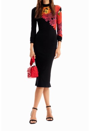 Kobieta w czarnej sukience midi z jasnymi kwiatowymi wzorami na ramieniu i rękawie, trzymająca czerwoną tłoczoną torebkę, ubrana w czarne szpilki.