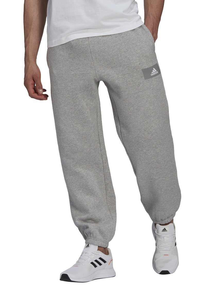 Šedé tepláky s elastickými manžetami, s malým šedým logem adidas na levém stehně. Nosené s bílými sportovními botami s černými akcenty.