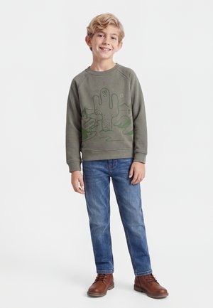 Garçon portant un sweat-shirt vert olive avec un motif cactus, jean bleu et bottes marron, debout et souriant sur fond blanc.