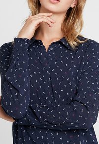 Blusa blu navy realizzata in tessuto morbido, caratterizzata da un motivo di piccole ancore bianche, rosa e viola, con collarino a bottoni e maniche lunghe.