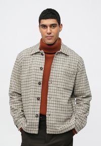 Muž oblečený v rustovom svetri s rolákom pod šedou a bielou károvanou zapínacou bundou, s rukami v vreckách na jednofarebnom pozadí.