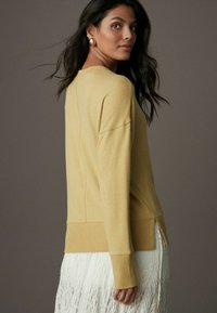 Pull en maille jaune avec poignets et ourlet côtelés, présentant des fentes sur les côtés. Le mannequin le porte par-dessus une jupe en dentelle blanche, mettant en valeur un look texturé.