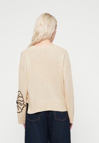 Béžový oversized svetr s žebrovaným lemem a manžetami, zobrazující černou květinovou výšivku na levém rukávu. Měkký texturovaný pletený materiál.