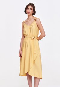 Żółta midi sukienka z miękkiego materiału, z regulowanymi ramiączkami z akcentami w formie pierścieni, z dopasowaną talią i dołem w stylu kopertowym.