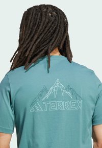 Teal tričkem s krátkým rukávem vyrobeným z měkké tkaniny. Na zadní straně má geometrický design hory a bílý potisk s nápisem "TERREX".