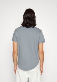 Joven con cabello oscuro a la altura de los hombros, vistiendo una camisa de manga corta gris claro y pantalones blancos, de espaldas contra un fondo sencillo.