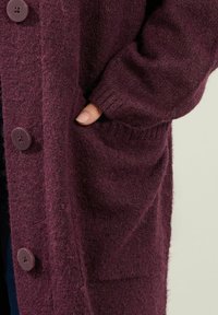 Lång, lila kofta i mjukt, strukturerat tyg med stora knappar och sidofickor. Koftan har en avslappnad passform och ribbade ärmslut.