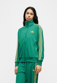 Νεαρή γυναίκα που φοράει πράσινη φόρμα Adidas με κίτρινες ρίγες και λογότυπο, στέκεται μπροστά σε απλό λευκό φόντο.