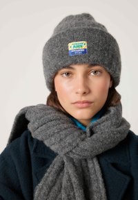 Noor inimene pruunide silmadega, kellel on hall kootud müts ja sobiv paks sall, sinine mantel, neutraalne ilme, hele taust.