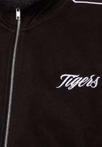Veste noire à fermeture éclair en tissu doux, avec des coutures blanches et un logo brodé "Tigers" sur le côté gauche de la poitrine.