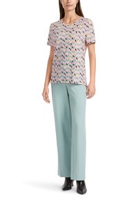 Mujer vestida con una blusa corta de manga en un patrón rosa, pantalones anchos de color azul claro y botines negros de tacón, de pie con una postura relajada.