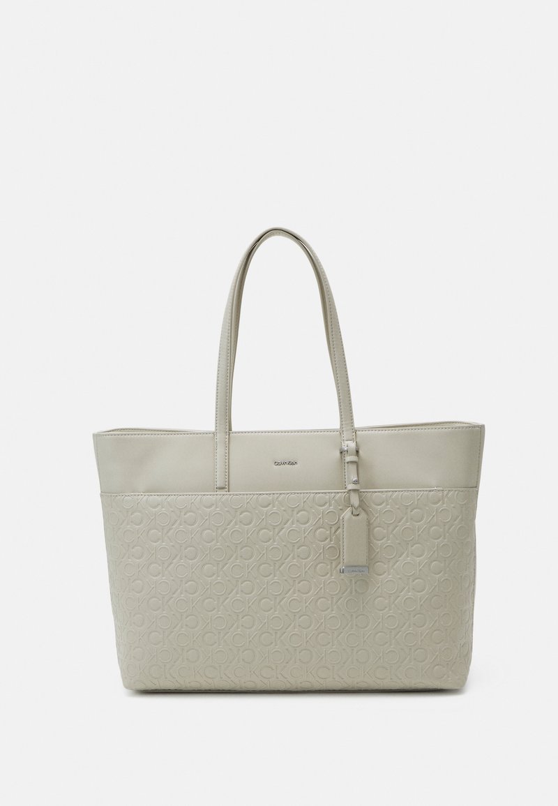 Sac fourre-tout beige Calvin Klein avec double poignée, motif de logo embossé sur la moitié inférieure et une étiquette suspendue à la poignée droite.