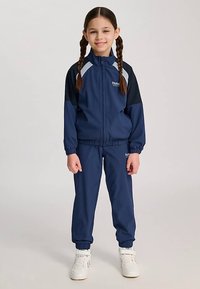 Marinblå träningsdräkt i lätt tyg med en zip-up jacka med svarta ärmar, vita detaljer samt elastiska mudd- och midjeband.