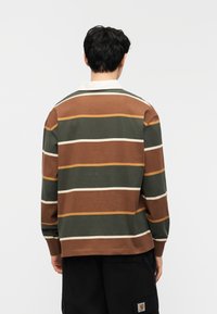 Άνδρας με την πλάτη, φορώντας μακρυμάνικο πουκάμισο σε καφέ, πράσινες και μπεζ ρίγες με λευκό γιακά και μαύρο παντελόνι cargo.