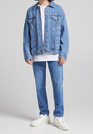 Person iført lyseblå denimjakke og matchende jeans med hvide sneakers og hvid T-shirt mod ensfarvet baggrund.