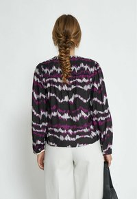 Camicia nera a maniche lunghe con motivi a onde viola e bianchi, caratterizzata da spalle arricciate e una vestibilità morbida. Abbinata a pantaloni chiari.