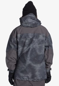 Vattentät grå jacka med mörkt abstrakt mönster, huva och elastiska ärmslut. Har ett lätt, texturerat tyg. Accessoarer visas inte.