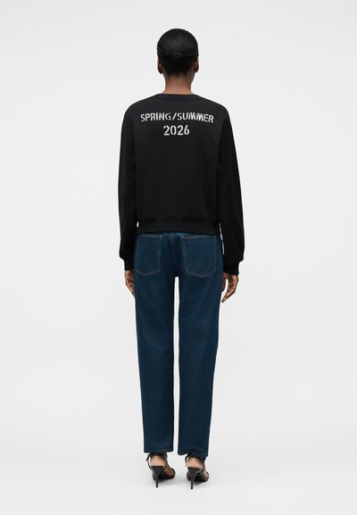 Άτομο που στέκεται με την πλάτη, φοράει μαύρο φούτερ με το κείμενο «SPRING/SUMMER 2026» στην πλάτη, μπλε τζιν και μαύρα παπούτσια με ψηλό τακούνι.