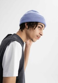 Lichtpaarse gebreide beanie op een persoon met gevlochten haar, gecombineerd met een grijze mouwloze trui over een wit T-shirt, gezien in profiel.