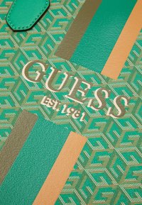 Materiale sintetico testurizzato verde e beige con un motivo geometrico, con il logo "GUESS" dorato e l'anno di fondazione 1981 in evidenza.