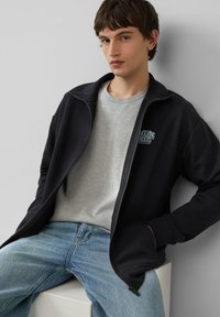 Zwarte zip-up jas over een grijs shirt, gecombineerd met blauwe jeans. Opmerkelijk logo detail op de jas. Gladde stof textuur, relaxte pasvorm.