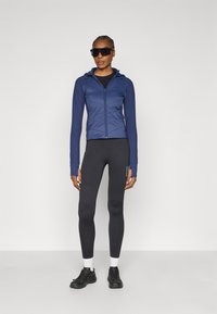 Tamsiai mėlynas sportinis švarkelis su užtrauktuku ir tekstūruota dalimi, derinamas su juodomis tamprėmis ir juodais sportiniais batais. Modelis dėvi tamsius saulės akinius.