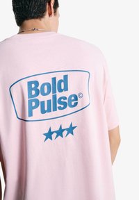 Roza oversized majica s bold modrim grafikom, ki na njej piše "Bold Pulse" in tremi modrimi zvezdami spodaj, izdelana iz mehke bombažne tkanine.