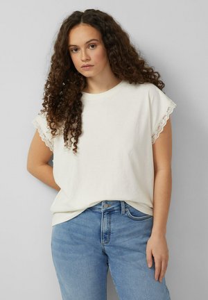 Junge Frau mit langen, lockigen Haaren, die ein weißes Kurzarm-Top mit Spitzenbesatz und blaue Jeans trägt, steht vor einer einfarbigen grauen Wand.