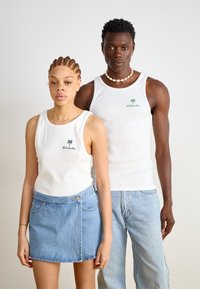Deux personnes portent des débardeurs blancs à côtes avec un logo de palmier vert. La femme porte une jupe enveloppante en denim, et l'homme l'associe avec un jean bleu clair.