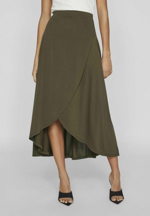 Femme portant une jupe portefeuille longue courte verte olive et des talons noirs ouverts au niveau des orteils, debout devant un fond gris clair uni.