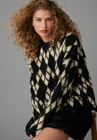 Pull en maille texturée à motif argyle noir et crème, avec une coupe décontractée et un col côtelé, associé à une jupe sombre.